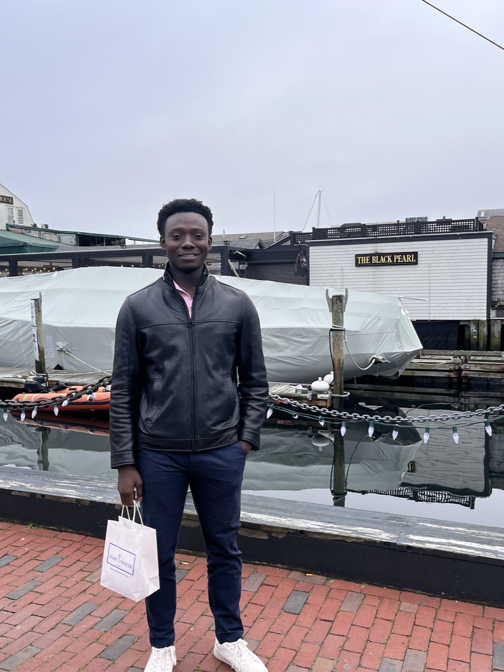 Standing on a wharf in a black leather jacket, holding a small paper shopping bag, a shrink-wrapped boat and the sign of The Black Pearl behind, under flat grey sky.