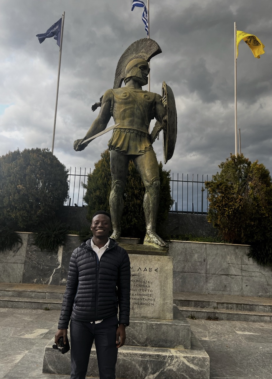 Standing at the foot of the bronze Leonidas statue in Sparta, its inscribed plinth and three flags behind, under heavy grey cloud.