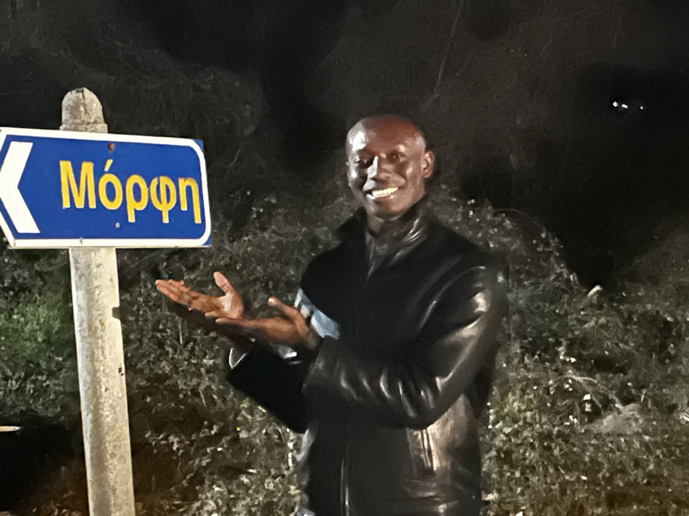 At night beside a blue Greek road sign reading Morphi, hands held open toward it, in a black leather jacket against a dark hedge.